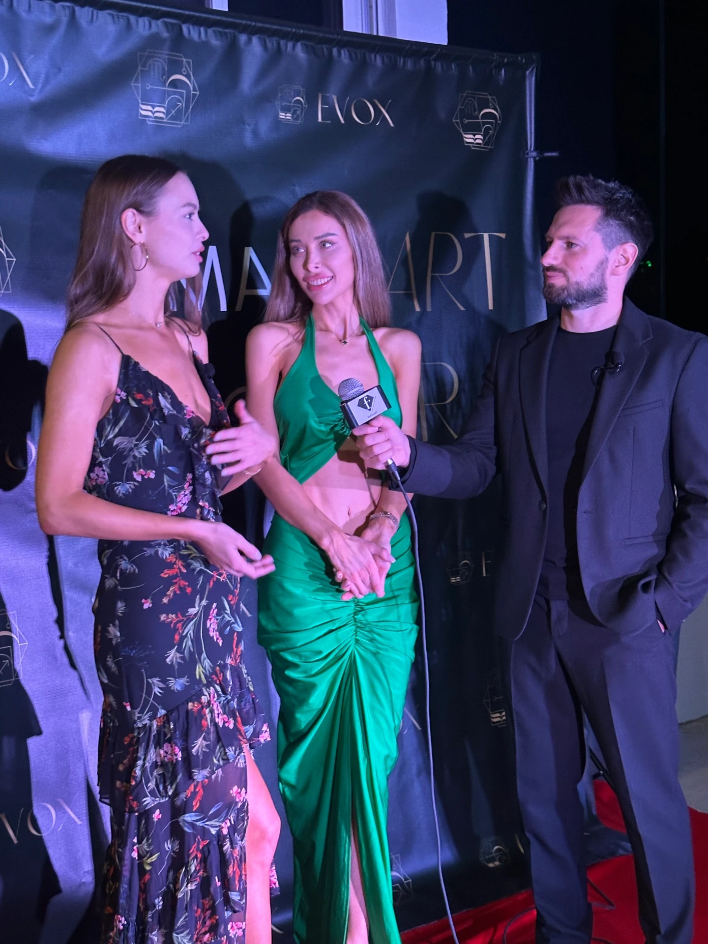Original event interview with two women and a man holding a Fashion TV microphone in front of the art-event backdrop